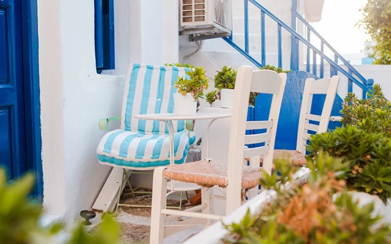 Blog 9 A small outdoor seating area with a whitewashed wall, blue trim, and a blue door. Two white chairs with blue cushions sit at a small white table. There are potted plants and greenery around the seating area.