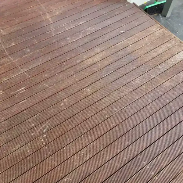 Decking Service 3 Wooden deck with dark brown planks adjacent to a white wall, showcasing marks and stains, with a green and black broom leaning nearby; ideal for cleaning or maintenance projects.