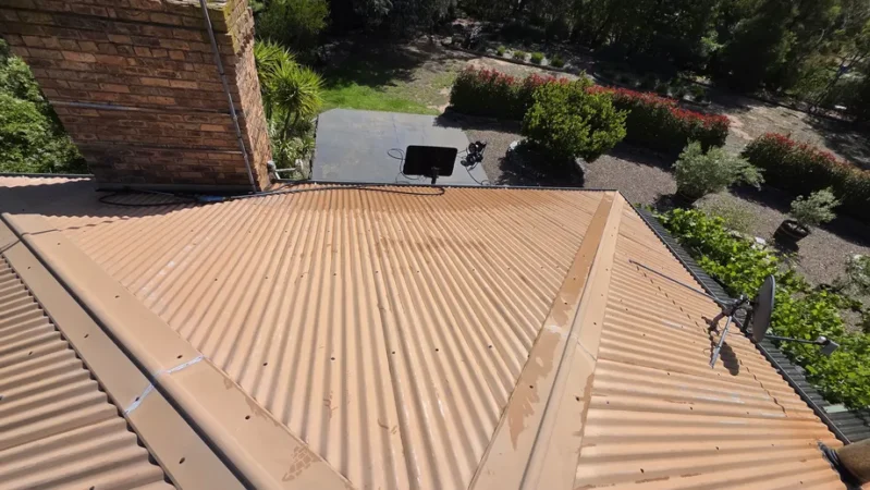 A faded tan corrugated roof surrounding a brick chimney, showing weathered surfaces before receiving professional roof painting and repair services in Bendigo.