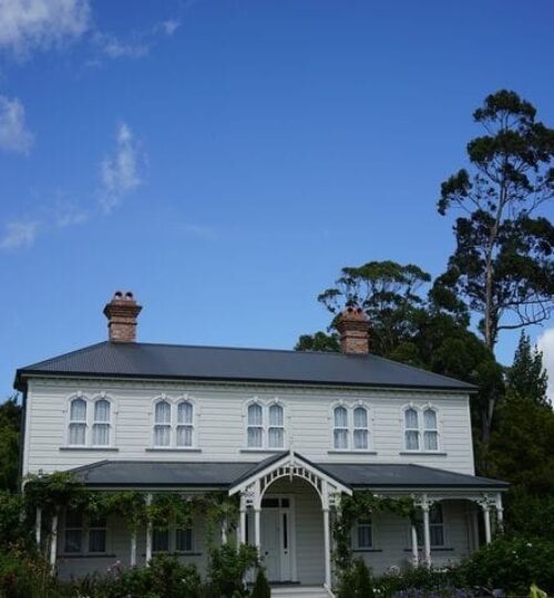 weatherboard house White house with three chimneys and a dark roof stands amidst lush greenery.